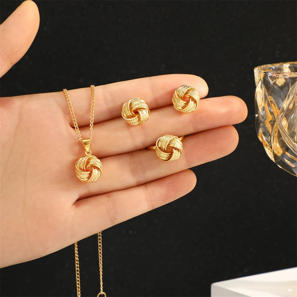 This jewelry set includes a necklace, earrings, and a ring made from shiny gold-colored alloy. The necklace features a delicate chain and a smooth twisted knot pendant. The lightweight earrings mimic this design, while the ring also showcases a twisted shape. Each piece offers a luxurious feel and is visually coordinated for an elegant look, displayed on soft satin fabric.