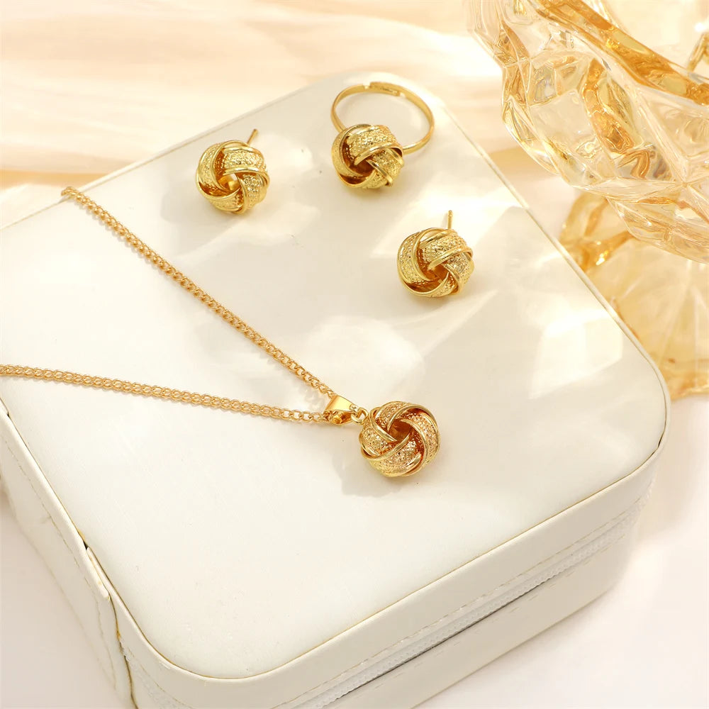 This jewelry set includes a necklace, earrings, and a ring made from shiny gold-colored alloy. The necklace features a delicate chain and a smooth twisted knot pendant. The lightweight earrings mimic this design, while the ring also showcases a twisted shape. Each piece offers a luxurious feel and is visually coordinated for an elegant look, displayed on soft satin fabric.