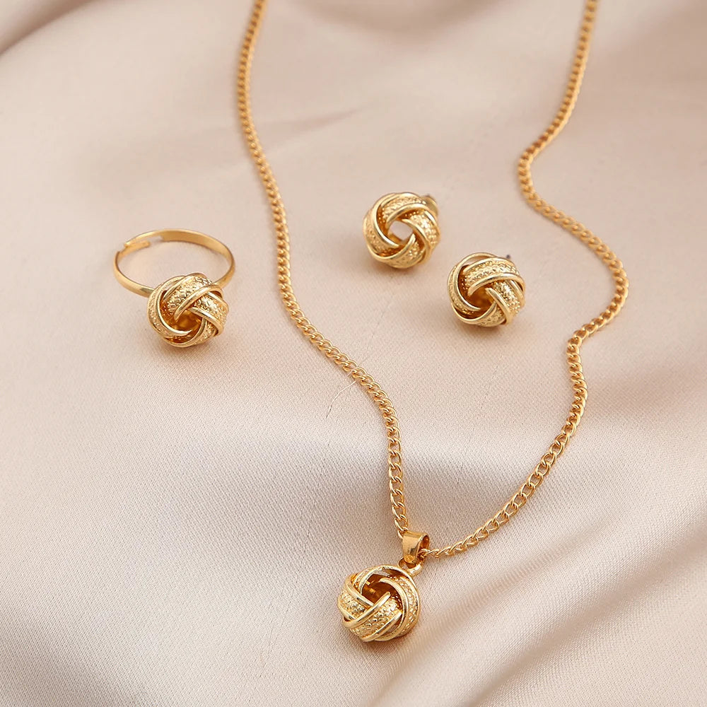 This jewelry set includes a necklace, earrings, and a ring made from shiny gold-colored alloy. The necklace features a delicate chain and a smooth twisted knot pendant. The lightweight earrings mimic this design, while the ring also showcases a twisted shape. Each piece offers a luxurious feel and is visually coordinated for an elegant look, displayed on soft satin fabric.