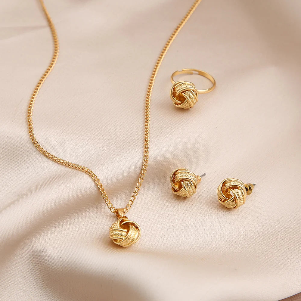 This jewelry set includes a necklace, earrings, and a ring made from shiny gold-colored alloy. The necklace features a delicate chain and a smooth twisted knot pendant. The lightweight earrings mimic this design, while the ring also showcases a twisted shape. Each piece offers a luxurious feel and is visually coordinated for an elegant look, displayed on soft satin fabric.