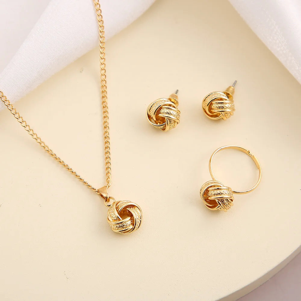 This jewelry set includes a necklace, earrings, and a ring made from shiny gold-colored alloy. The necklace features a delicate chain and a smooth twisted knot pendant. The lightweight earrings mimic this design, while the ring also showcases a twisted shape. Each piece offers a luxurious feel and is visually coordinated for an elegant look, displayed on soft satin fabric.